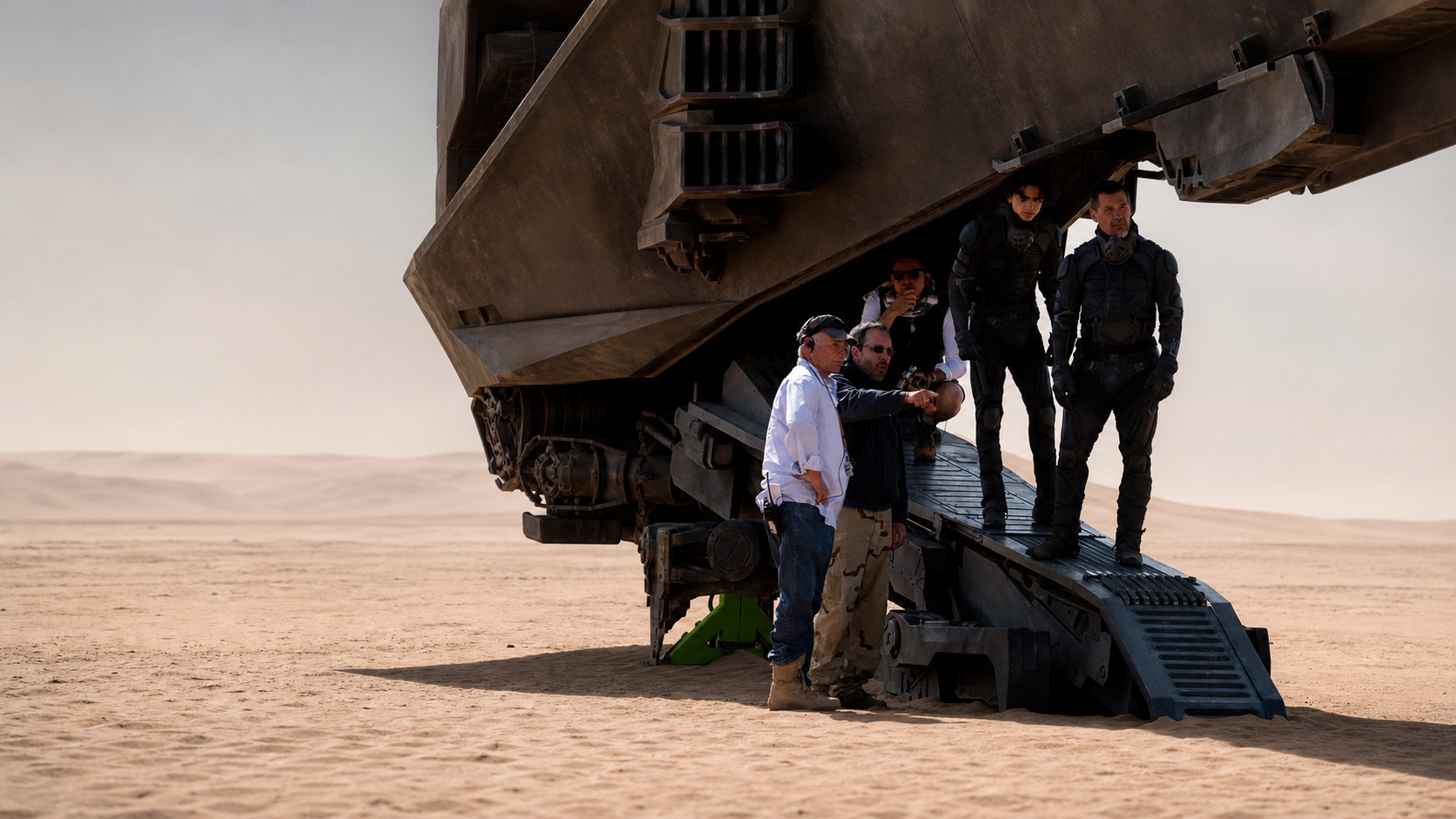 A widescreen Cinema One production-watch visual derived from a real Dune behind-the-scenes desert photograph, with crew and costumed performers beneath a practical aircraft set.